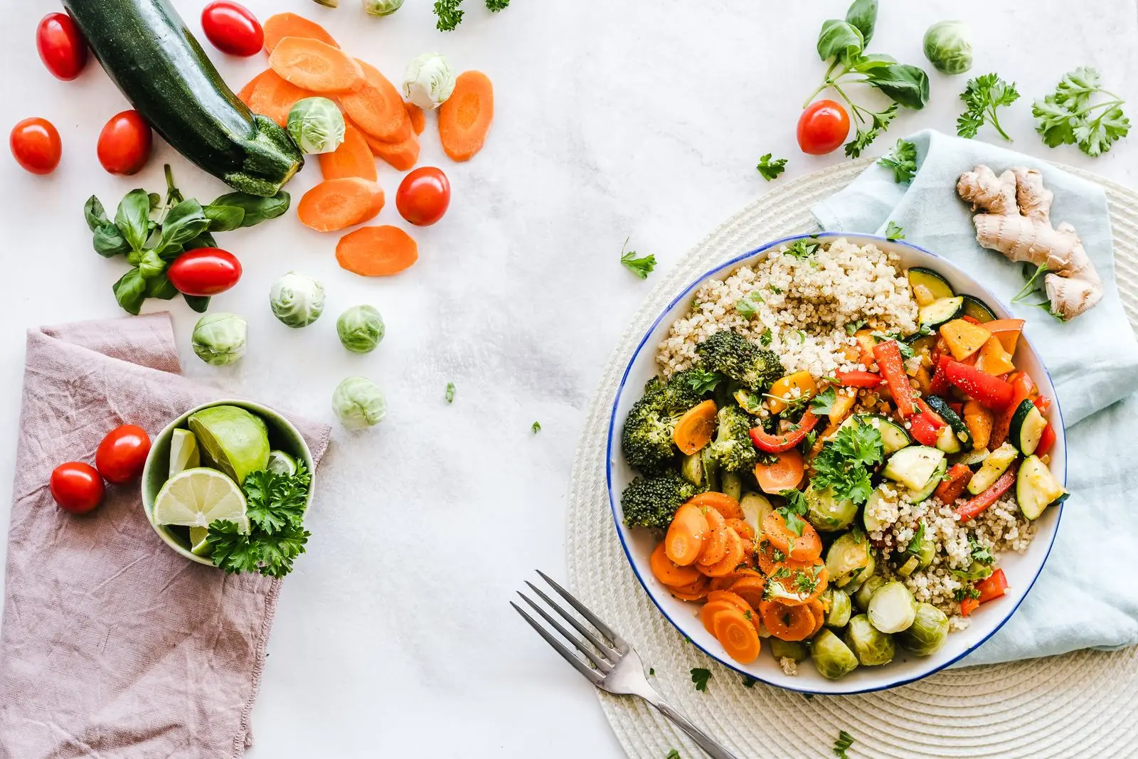 A colourful buddha bowl of quinoa and roasted vegetables beside fresh ingredients on a marble surface.