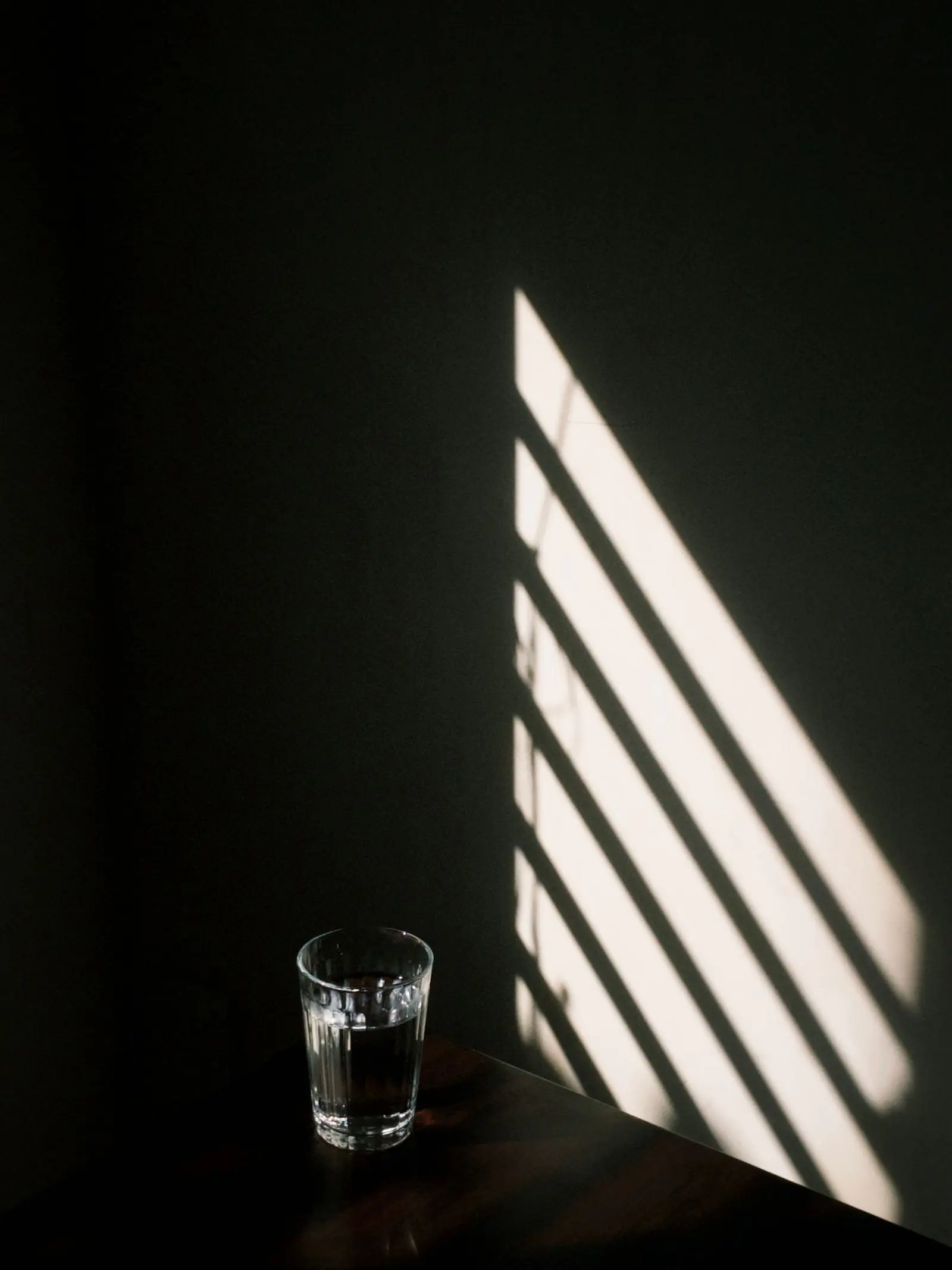 A glass of water on a dark table beneath the striped shadow of window blinds.