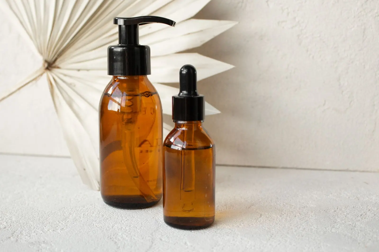 An amber pump bottle and a dropper bottle standing beside a dried decorative palm frond.